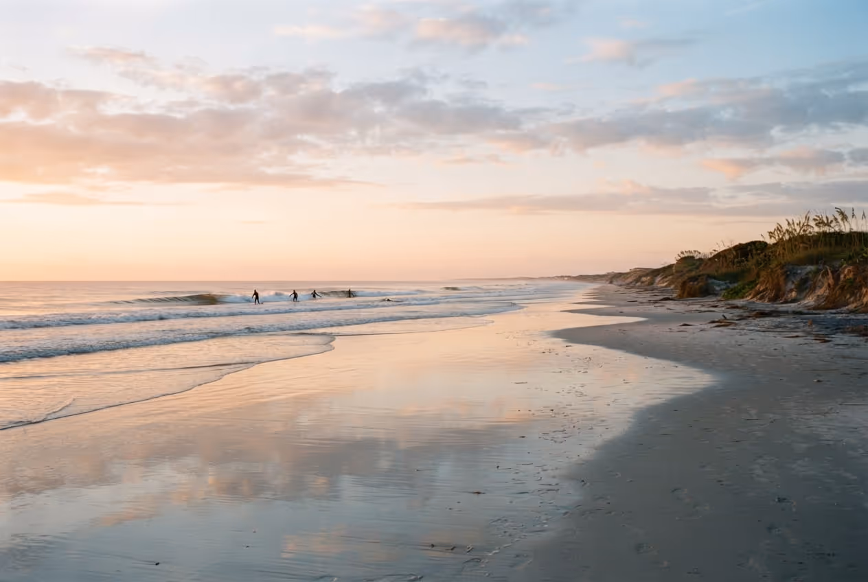 Faixa larga de areia compacta em New Smyrna Beach na maré baixa, com surfistas pequenos ao longe e dunas à direita